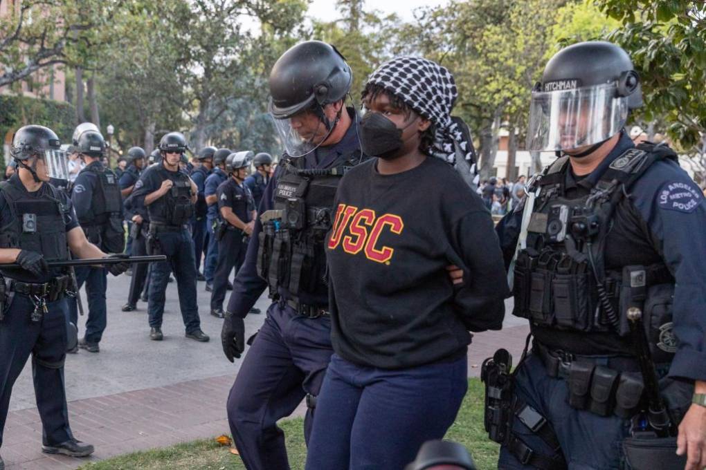 A Black female student in a headscarf and 'USC' sweatshirt is led away with her hand behind her back by police in riot gear.