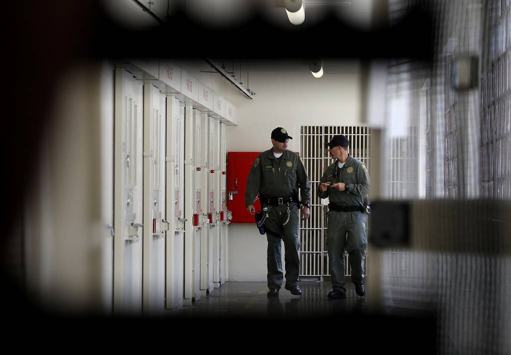 The hallway of a prison with two guards seen through the frame of bars.