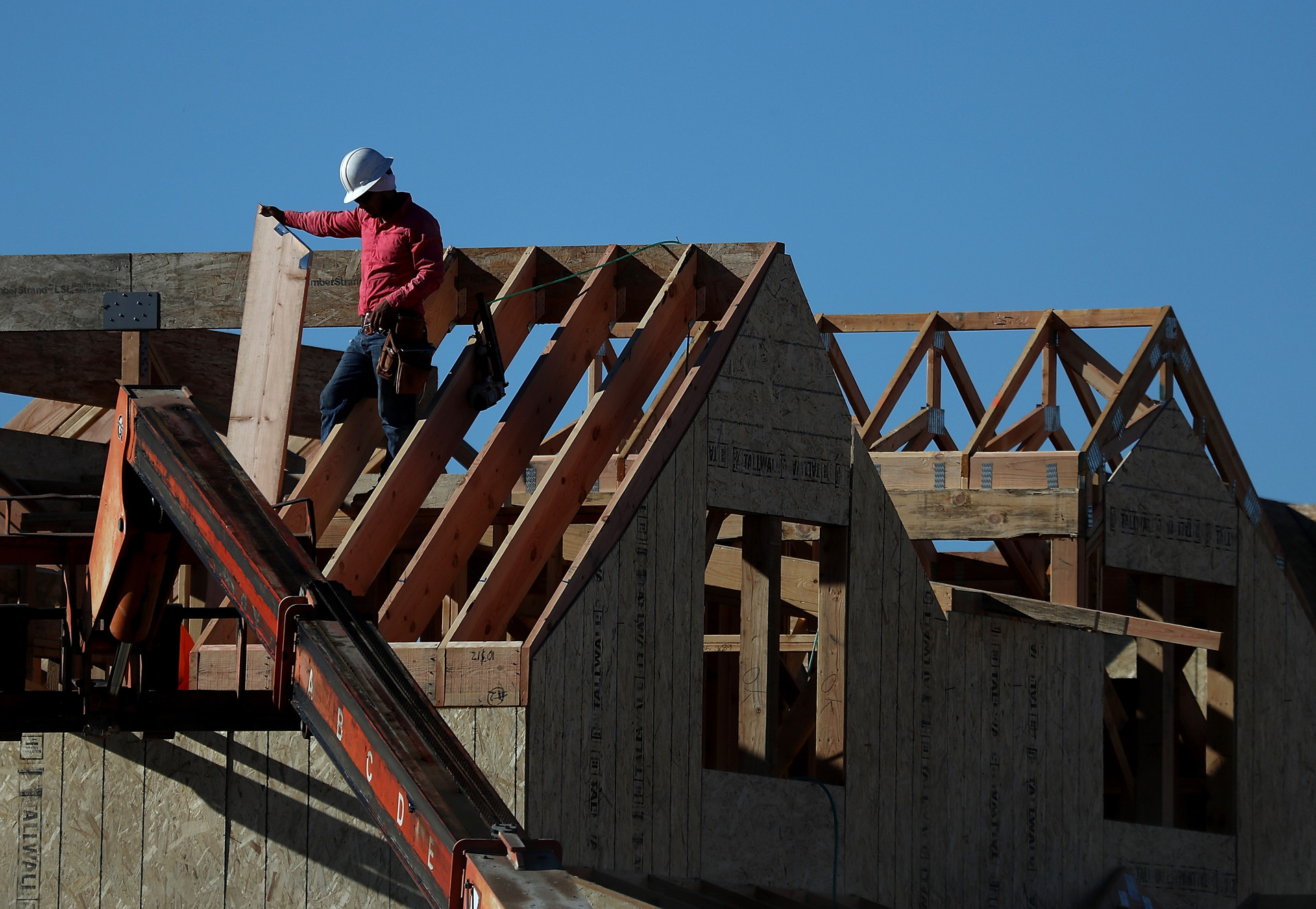 A person wearing construction equipment holds a wooden support beam while a roof is built.