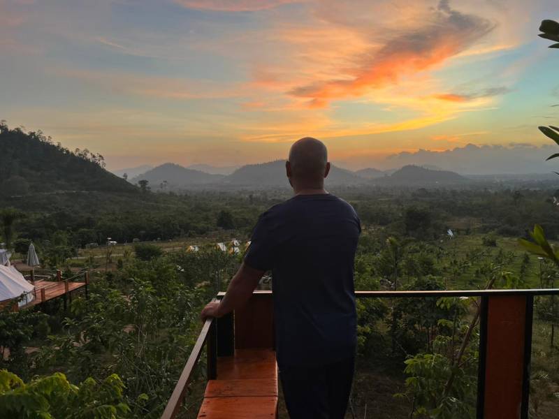 A man seen from behind stands on a balcony at sunset, looking out at a lush forest and mountain landscape.