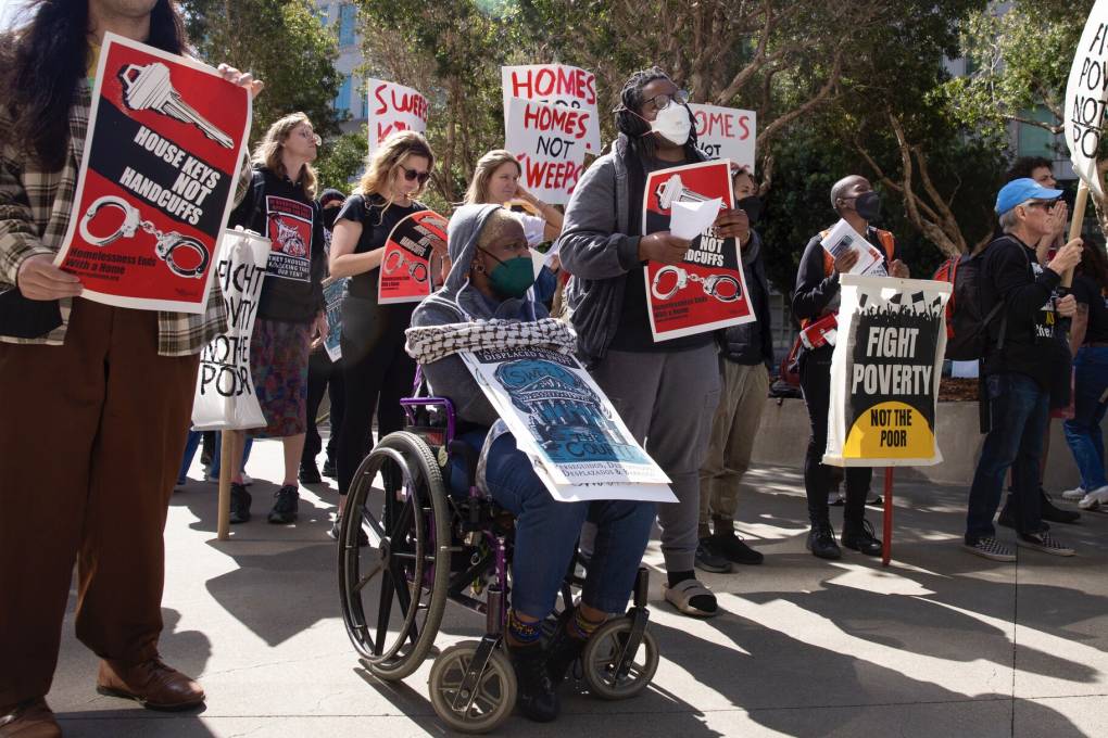 A Black woman in a wheelchair is among a group of protesters holding signs like 'Fight Poverty' and 'Homes not Sweeps.'