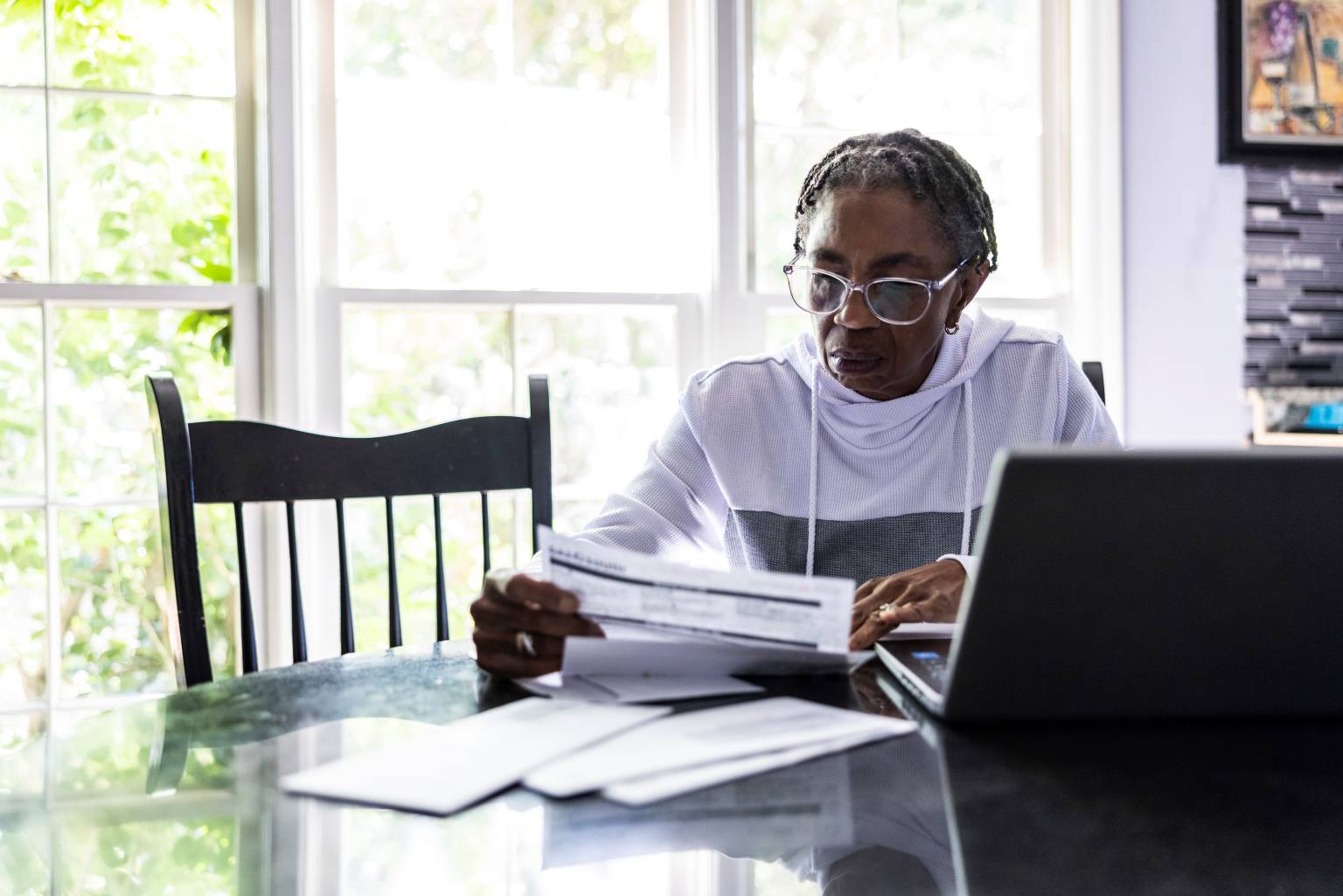 Una persona mayor de edad está sentada en una mesa con una computadora y varios papeles frente a ella.
