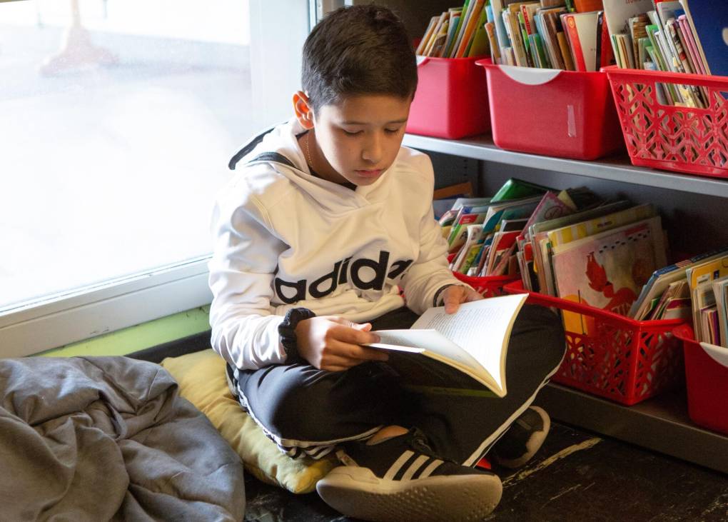 A young boy wearing a white hoodie and black pants reads a book while sitting down.