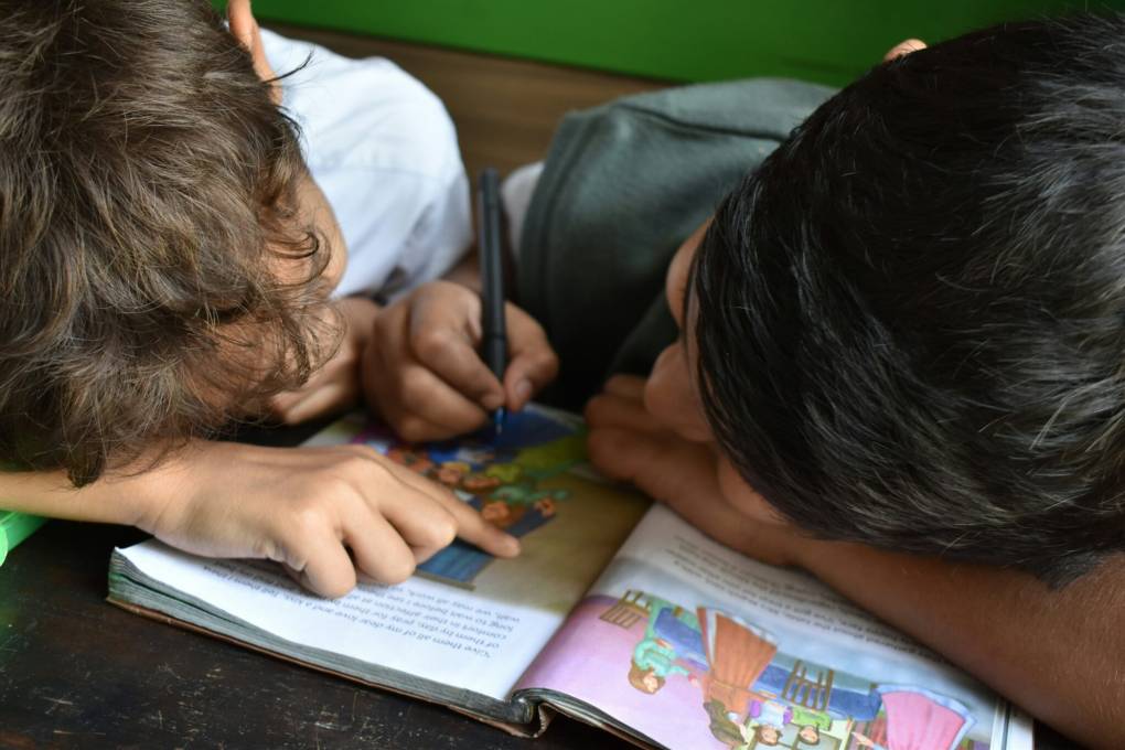 Two children rest their heads on an open book with one child holding a writing instrument.