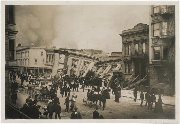 Black and white photo of nearly flattened buildings, with people walking by on the street.