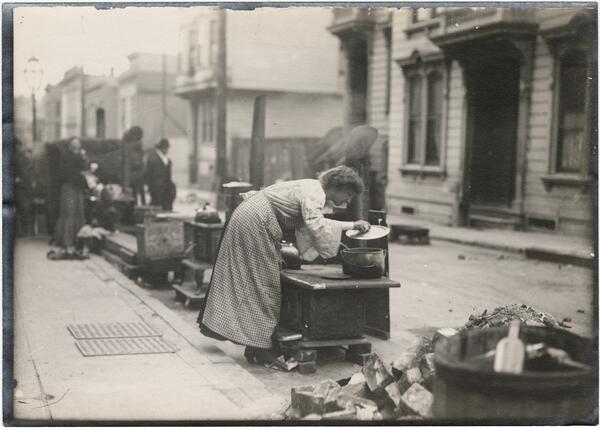 Black and white photo of of a woman cooking on a cast iron stove in the street.