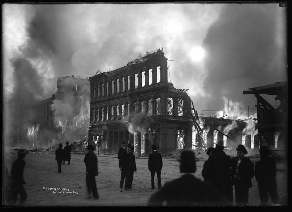 Dramatic black and white photo of a fierce fire burning behind the remains of a building.