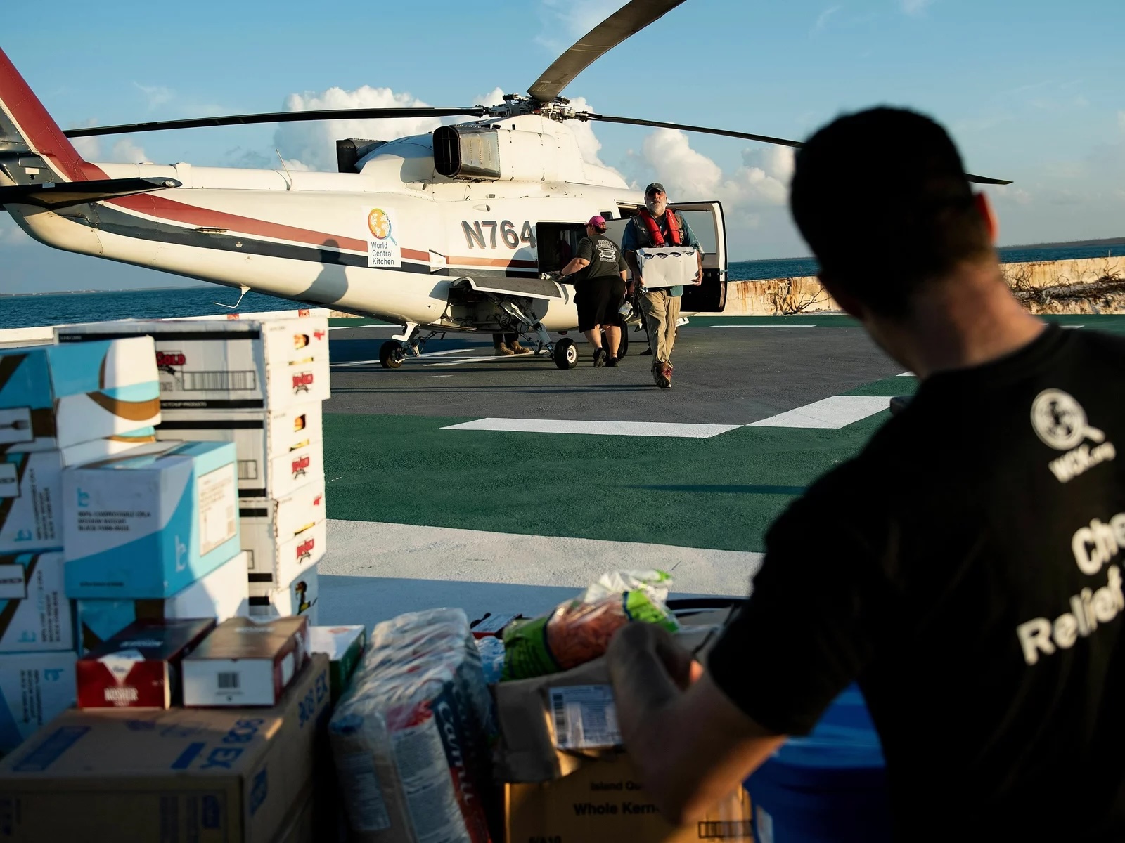 A white helicopter on a tarmac with people taking cases and boxes out of it, and another peron in foreground arranging boxes of food and supplies.