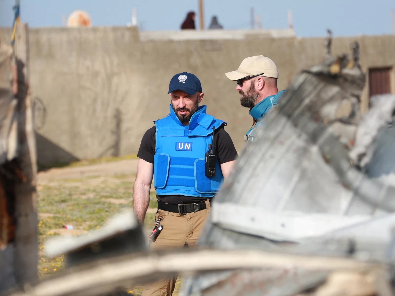 Two men with hats and blue vests speak behind a wreckage.