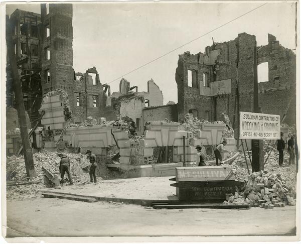 Black and white photo of two men shoveling debris in front of burned out buildings.