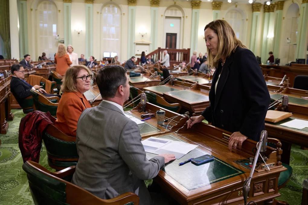 Assemblymember Jacqui Irwin stands over two colleagues who are sitting behind desks.