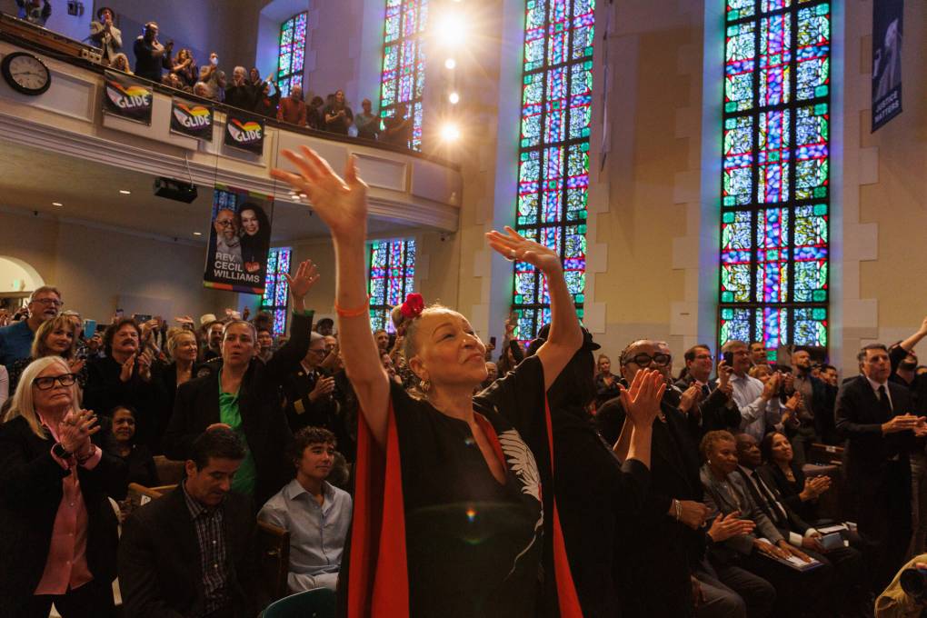 A woman with arms raised dancing stands in a church with lights and stained glass behind her.