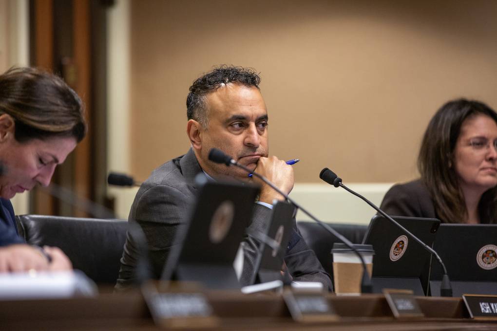 Two women and a man sit behind desks with microphones and name placards in a room.