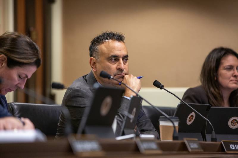 Two women and a man sit behind desks with microphones and name placards in a room.