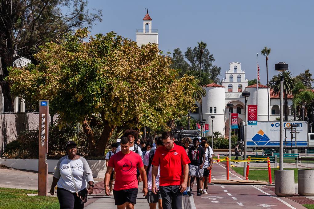 People walk down a street with a college campus in the background.