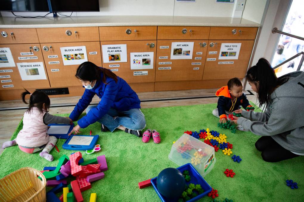 Two mothers play with their young children on a green rug inside a classroom.