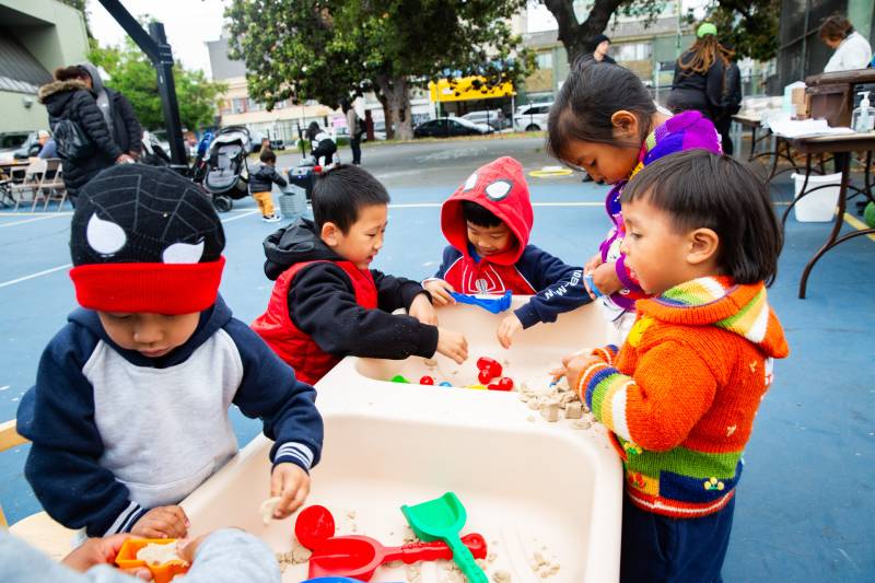 Several young children play with toys on tables on a court outside.