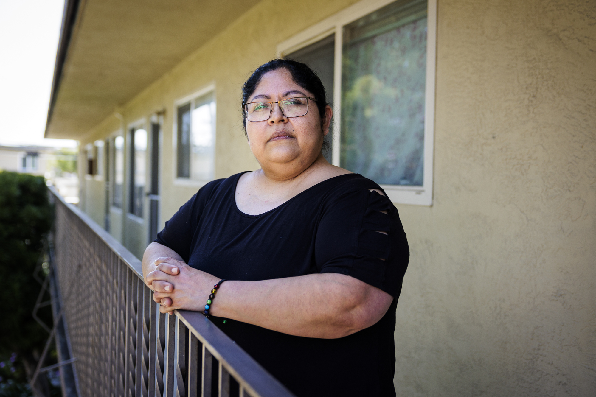 A Latina woman wearing glasses and a black short-sleeved shirt stands on the balcony of an apartment building with her hands resting on the rails.