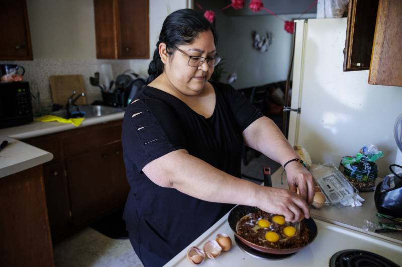 A Latina woman wearing glasses and a black short-sleeve shirt stands in front of a kitchen stove cooking eggs.