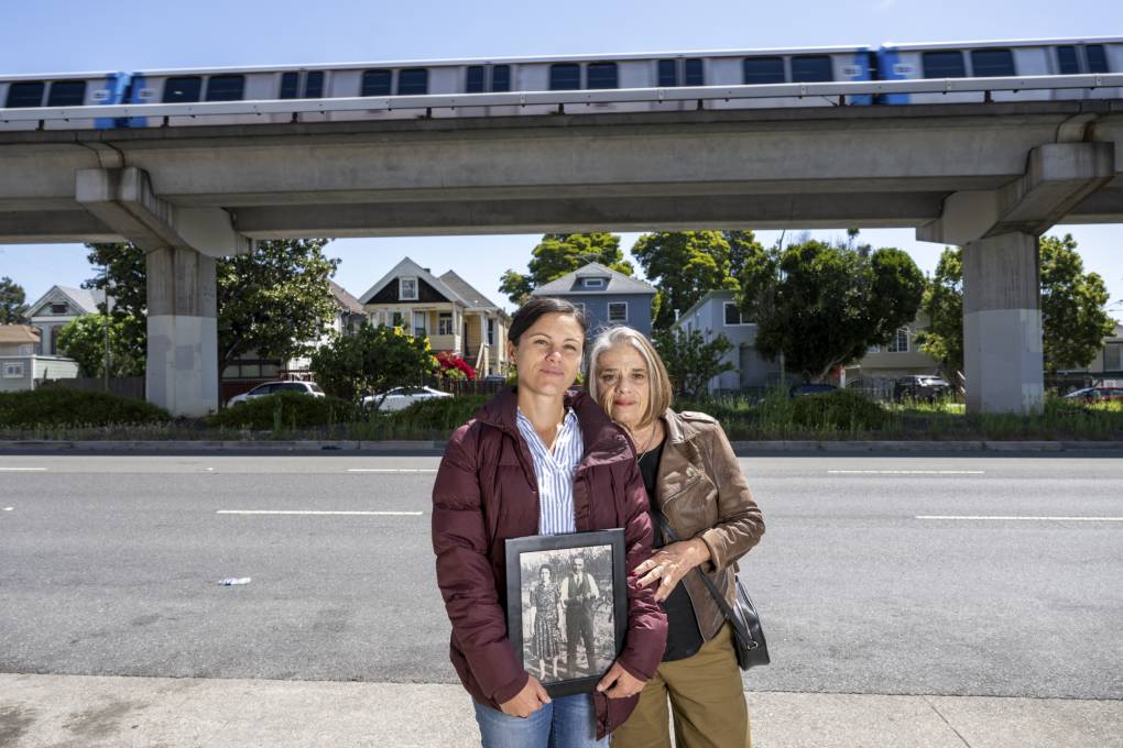 A younger woman and an older woman stand in front of a train track, holding a black and white photo.