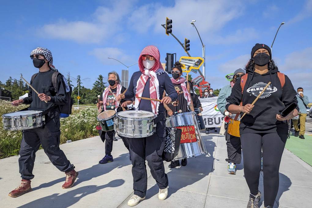 A group of people wearing masks and holding drums walk down the street.