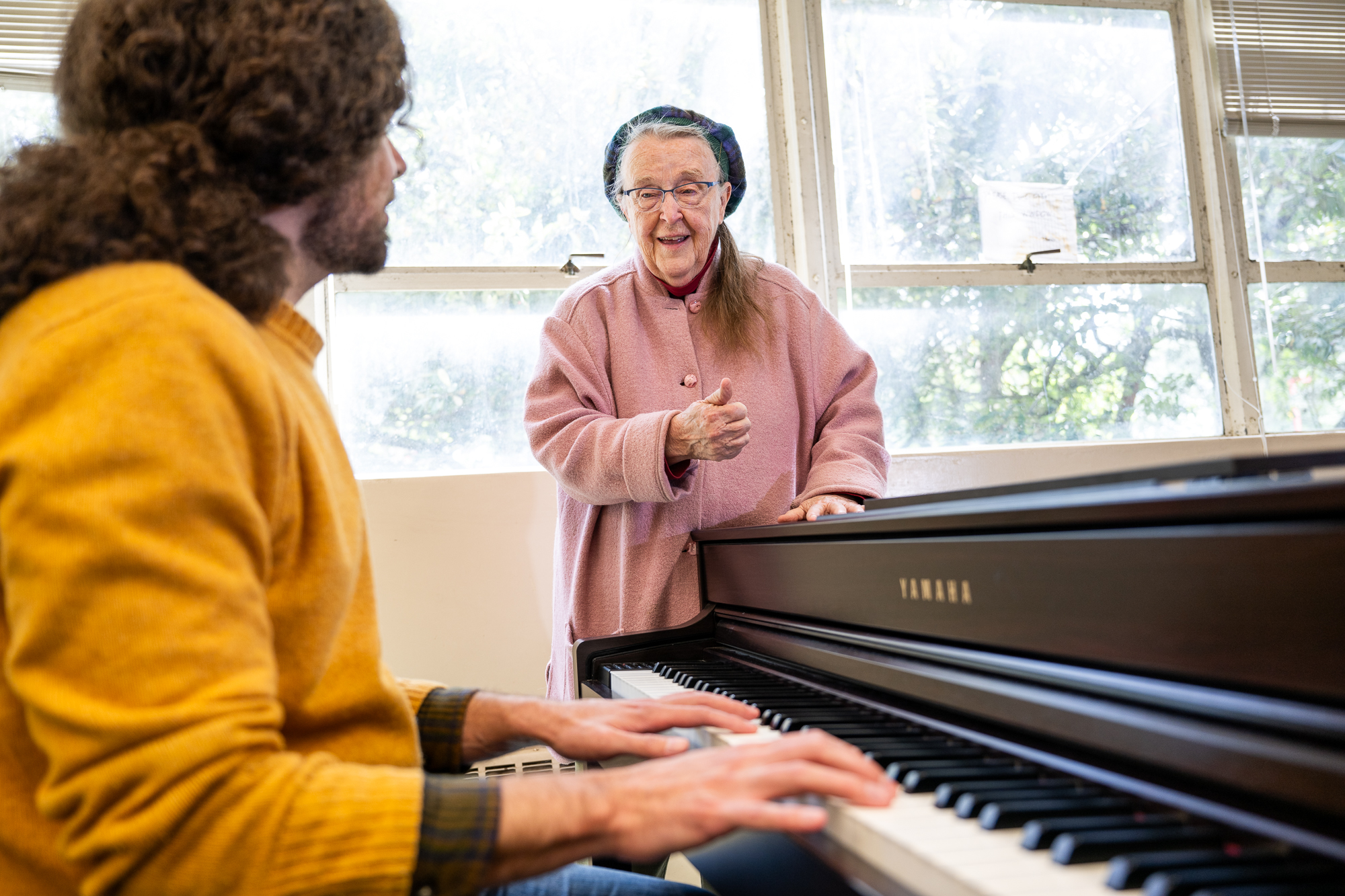 An older woman wearing glasses and a pink coat speaks to a younger student as he plays a grand piano