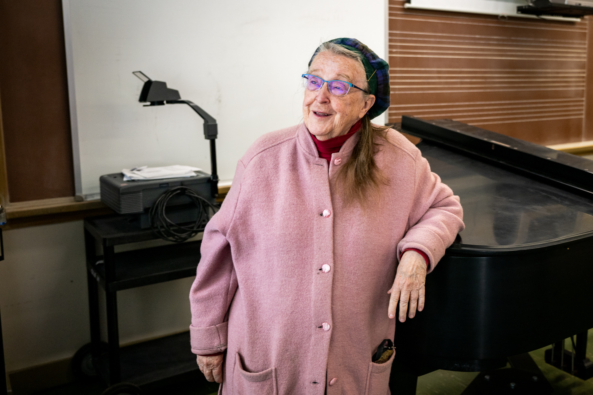 An older woman wearing glasses and a pink coat smiles as she leans on a grand piano