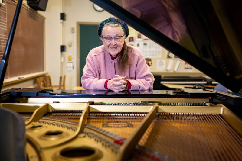 An older woman wearing glasses and a pink coat smiles as she leans on a grand piano