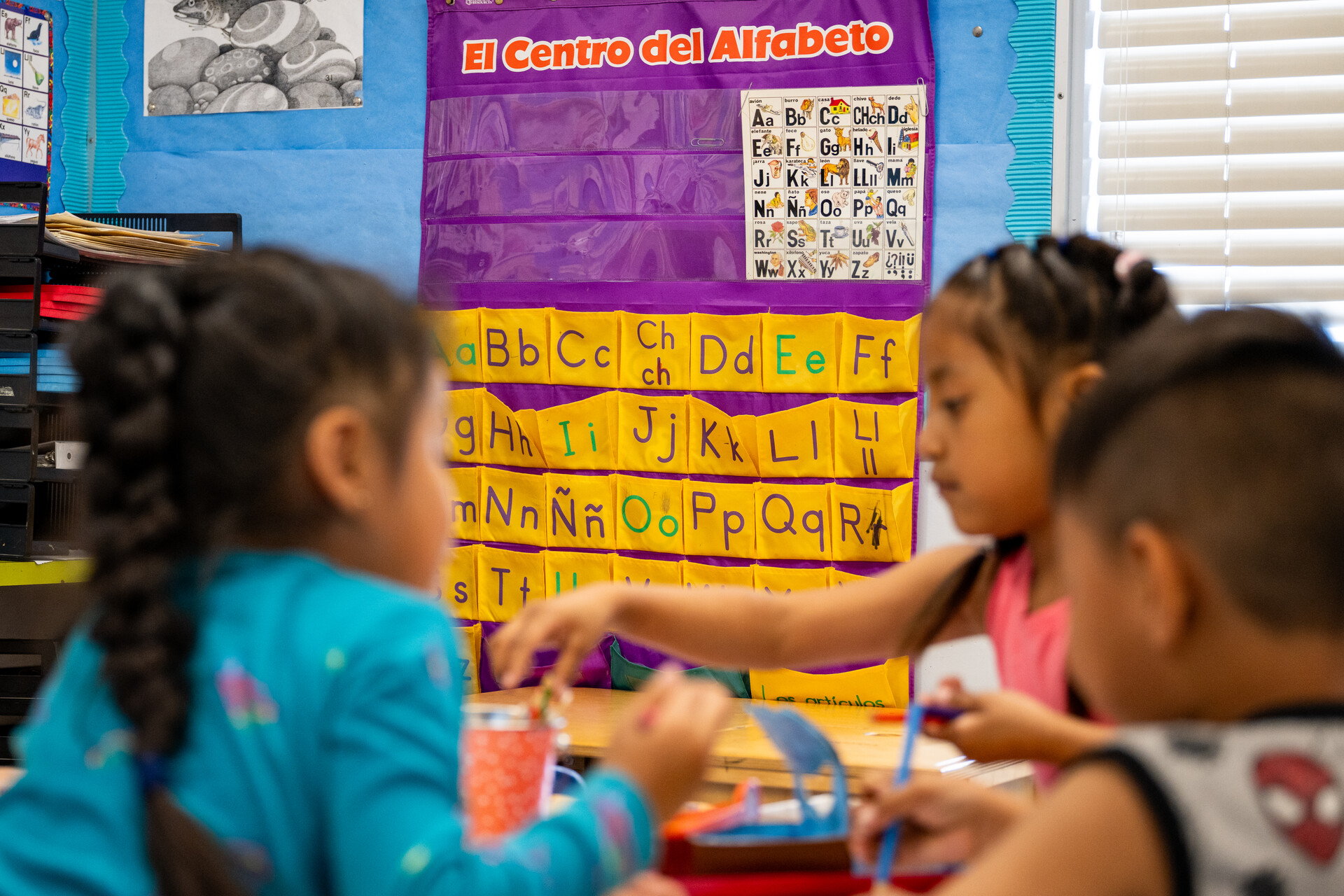 Three young children in the foreground work on an exercise as a bilingual alphabet hangs on the wall of a classroom behind them