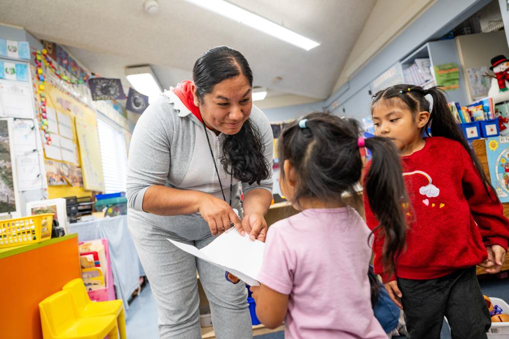 A woman teacher leans over and smiles at two young students in a classroom.