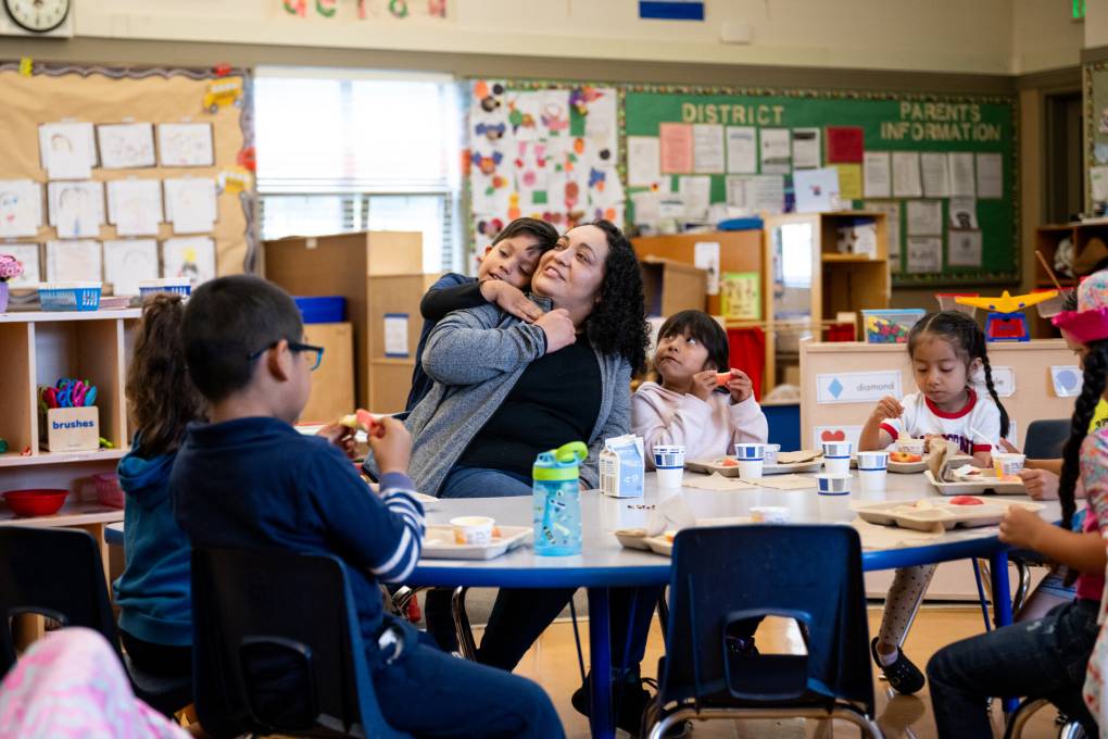 A teacher sits at a classroom table with young students as one student puts his arm tenderly around her neck