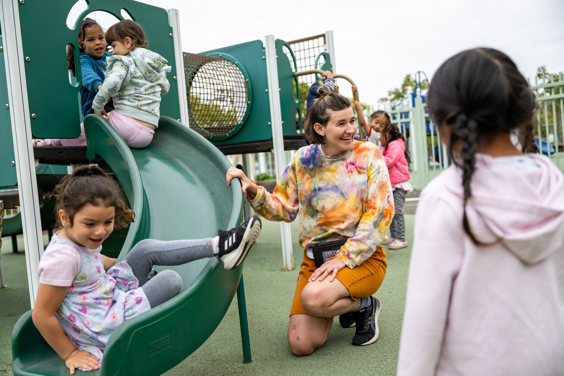 A teacher smiles as she plays with students at an outdoor play gym slide