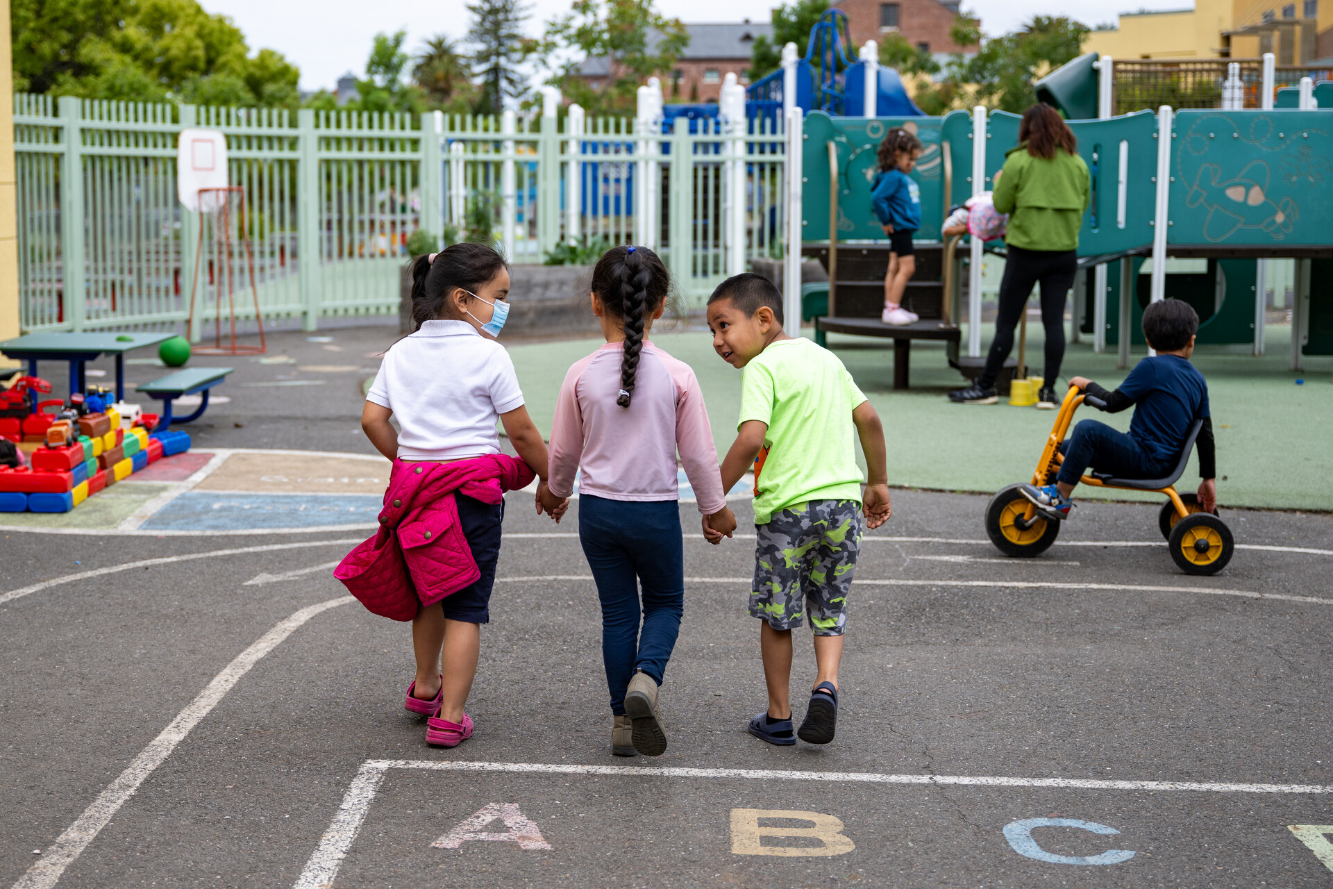 Three young students hold hands outside as they walk away from the camera towards a play gym structure during recess