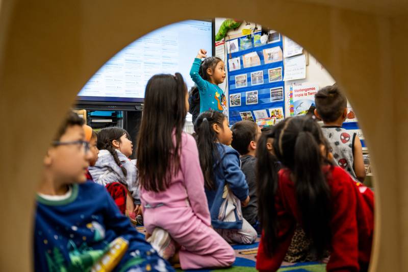 A young student stands and raises her hand in class as other students around her remain seated in a classroom