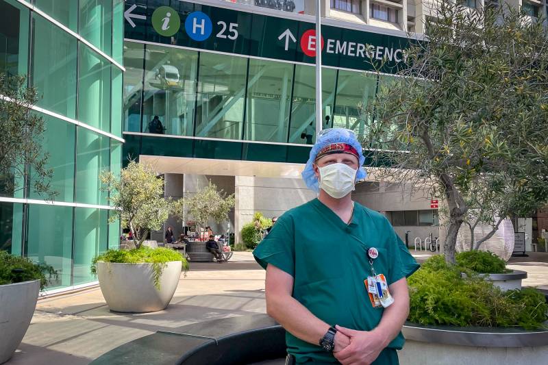 A man wearing a surgical facemask and green scrubs stands outside a hospital building