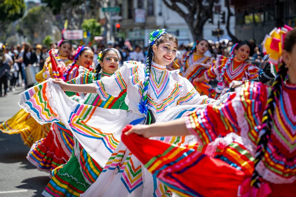 Several women dressed in colorful clothing dance and walk down the street during a parade.