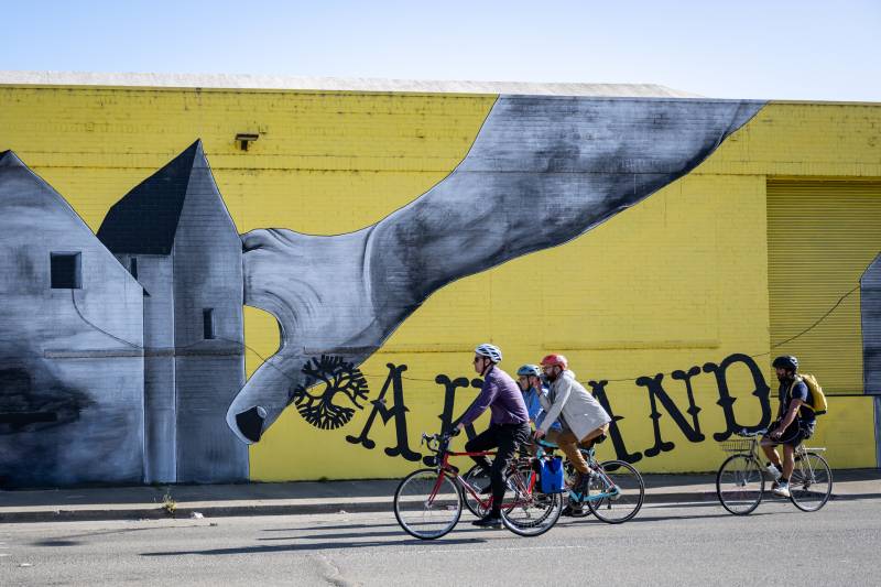 A group of cyclists ride bicycles in the foreground with a large, graphic yellow painting on a building appears in the background, with a hand reaching down and grasping a building with the word 'Oakland' emblazoned over it.