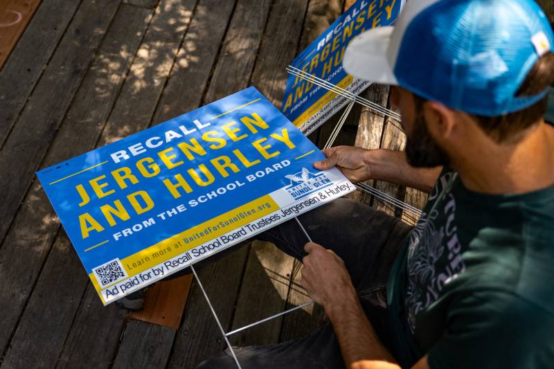 A bearded man sits outside while assembling blue and yellow yard signs.