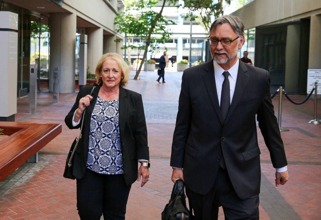 A middle-aged white woman with blond hair walks through a courtyard, next to a middle-aged man wearing a suit and carrying a briefcase.