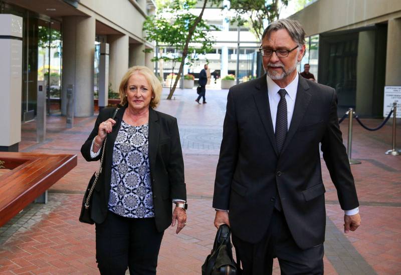 A middle-aged white woman with blond hair walks through a courtyard next to a middle-aged man wearing a suit and carrying a briefcase.