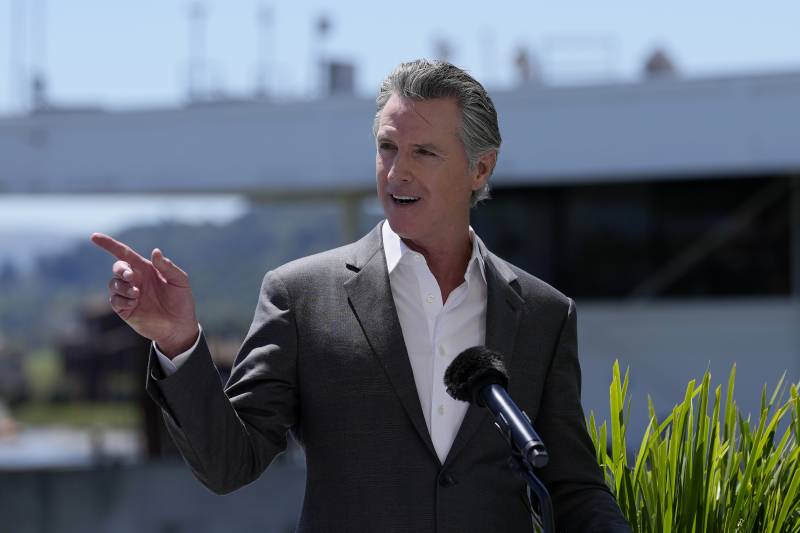 A white, middle-aged man in a suit and white shirt with no tie gestures to his right while speaking into a microphone outdoors.