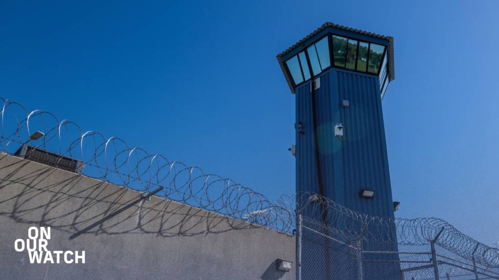 Image of a prison guard tower. In the foreground, a gray concrete wall topped with barbed wire transitions into a chain-link fence on the right, also crowned with barbed wire. In the right third of the frame, a prominent five-sided dark blue guard tower ascends into a cloudless deep blue sky. The top of the tower widens into a control room covered with reflective windows.