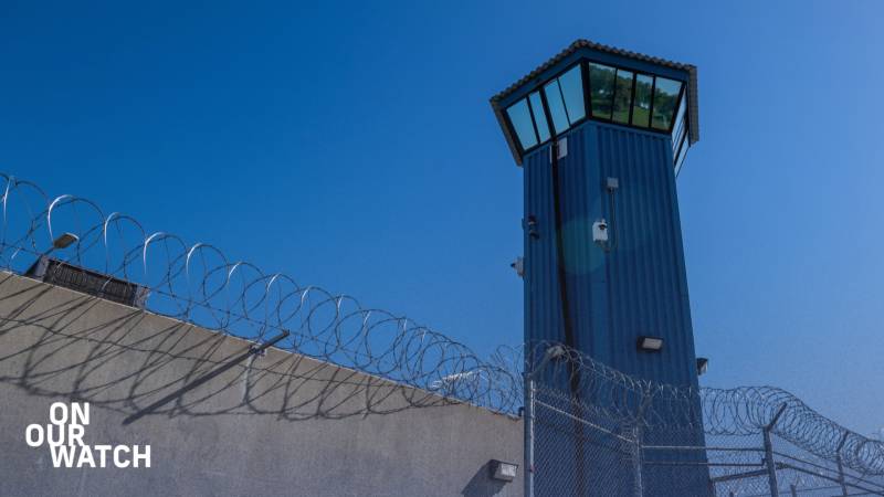 Image of a prison guard tower. In the foreground, a gray concrete wall topped with barbed wire transitions into a chain-link fence on the right, also crowned with barbed wire. In the right third of the frame, a prominent five-sided dark blue guard tower ascends into a cloudless deep blue sky. The top of the tower widens into a control room covered with reflective windows.