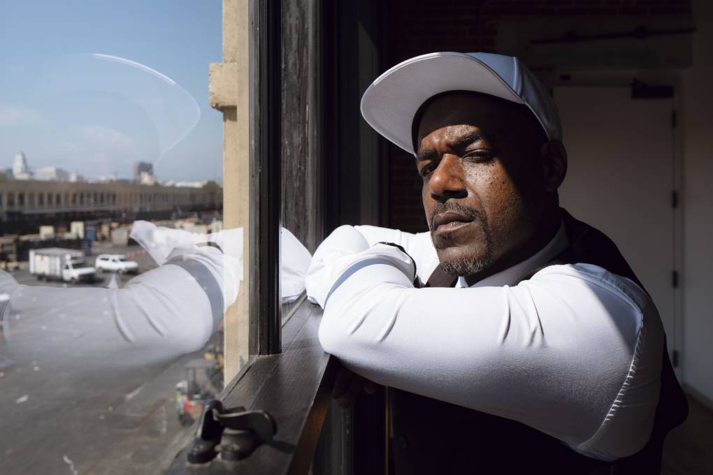 A Black man wearing a cap and white shirt rests his arms and looks out a window.