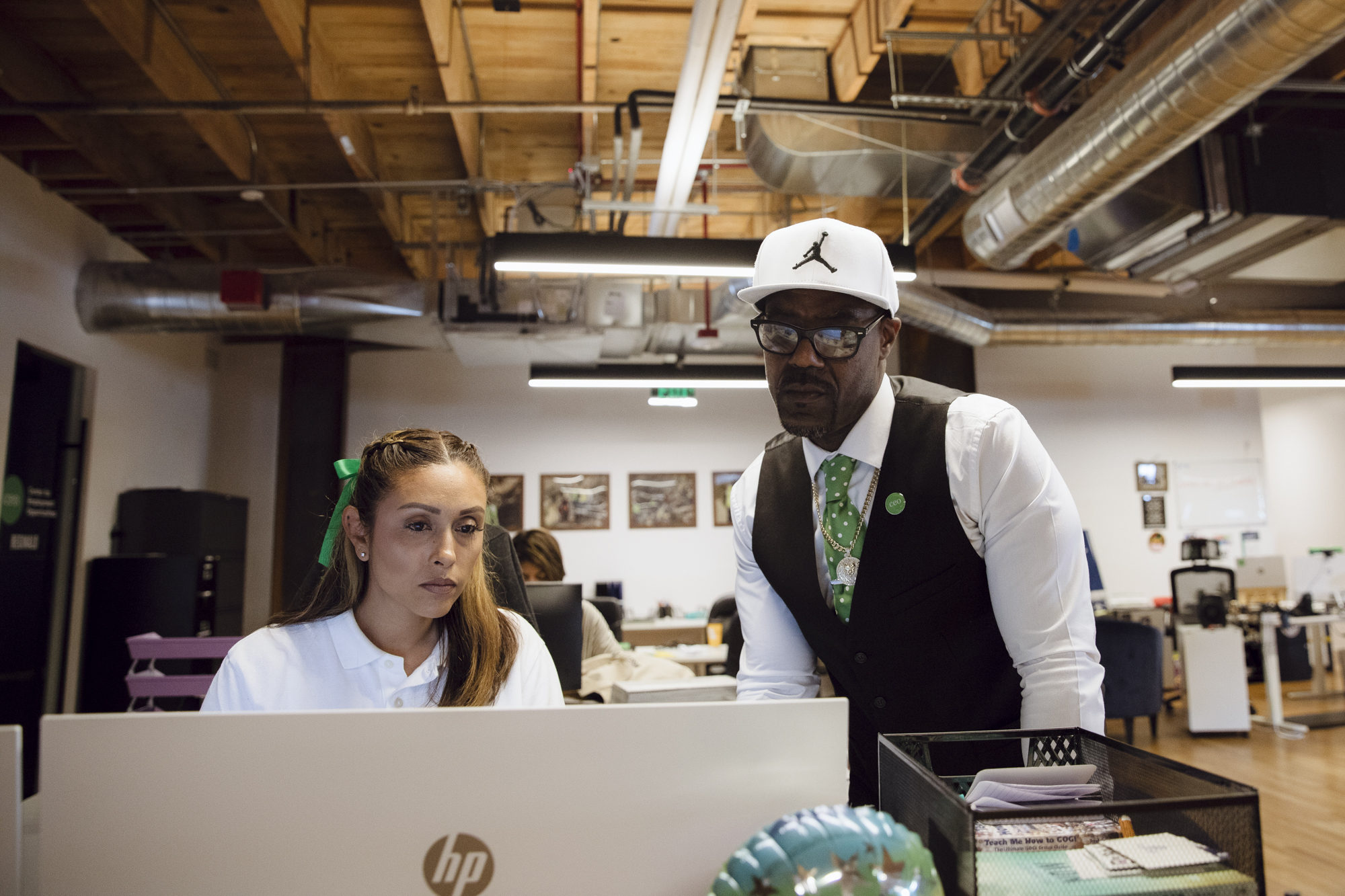 A Black man wearing a white hat, black vest and white shirt stands over a woman wearing a white shirt looking at a desktop screen in a building.