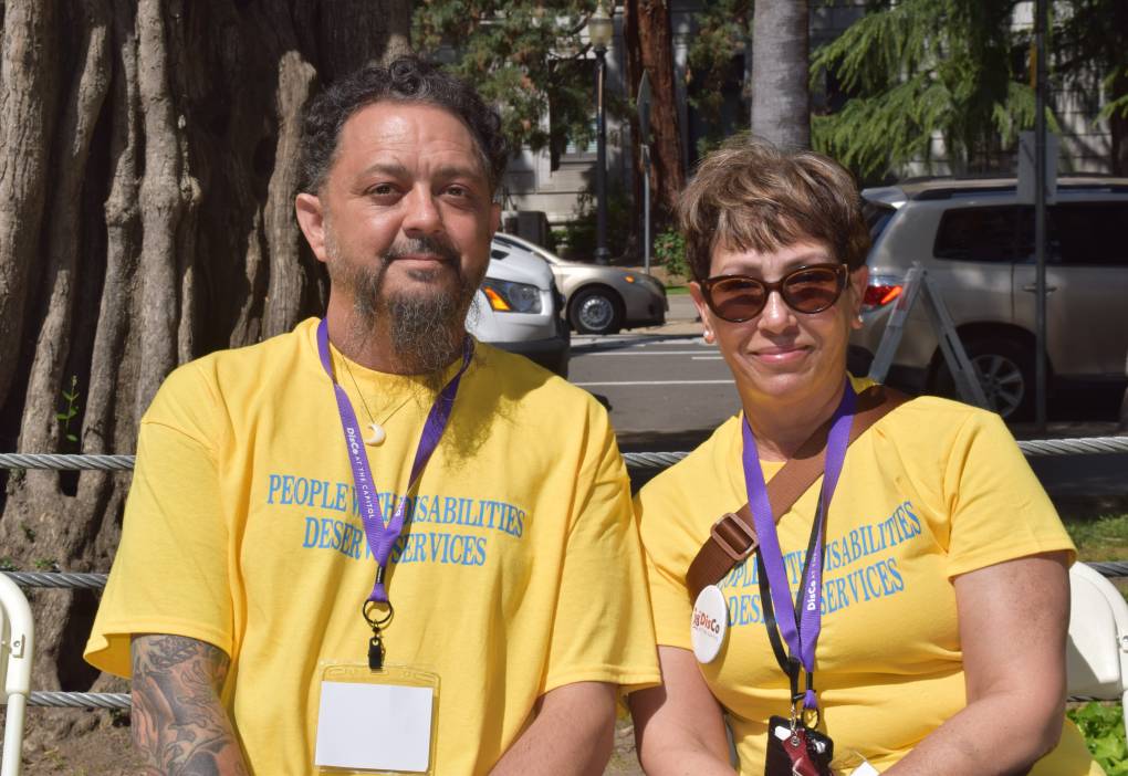 A man and woman wearing yellow shirts with purple lanyards sit next to each other outside.