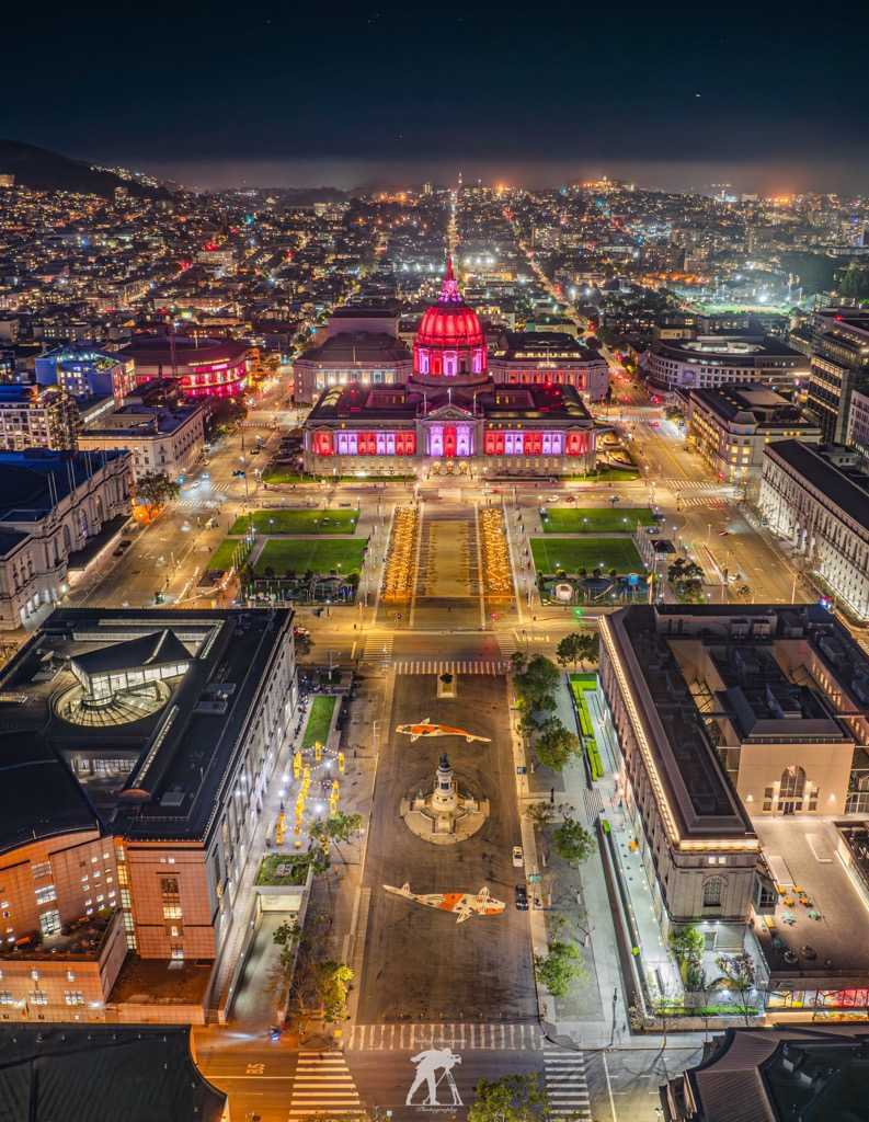 An aerial image of a pedestrian plaza, where two large koi fish "swim" around a statue. It's night time and the city lights are on display in surrounding buildings. A block away, San Francisco City Hall is illuminated with red white and blue. 