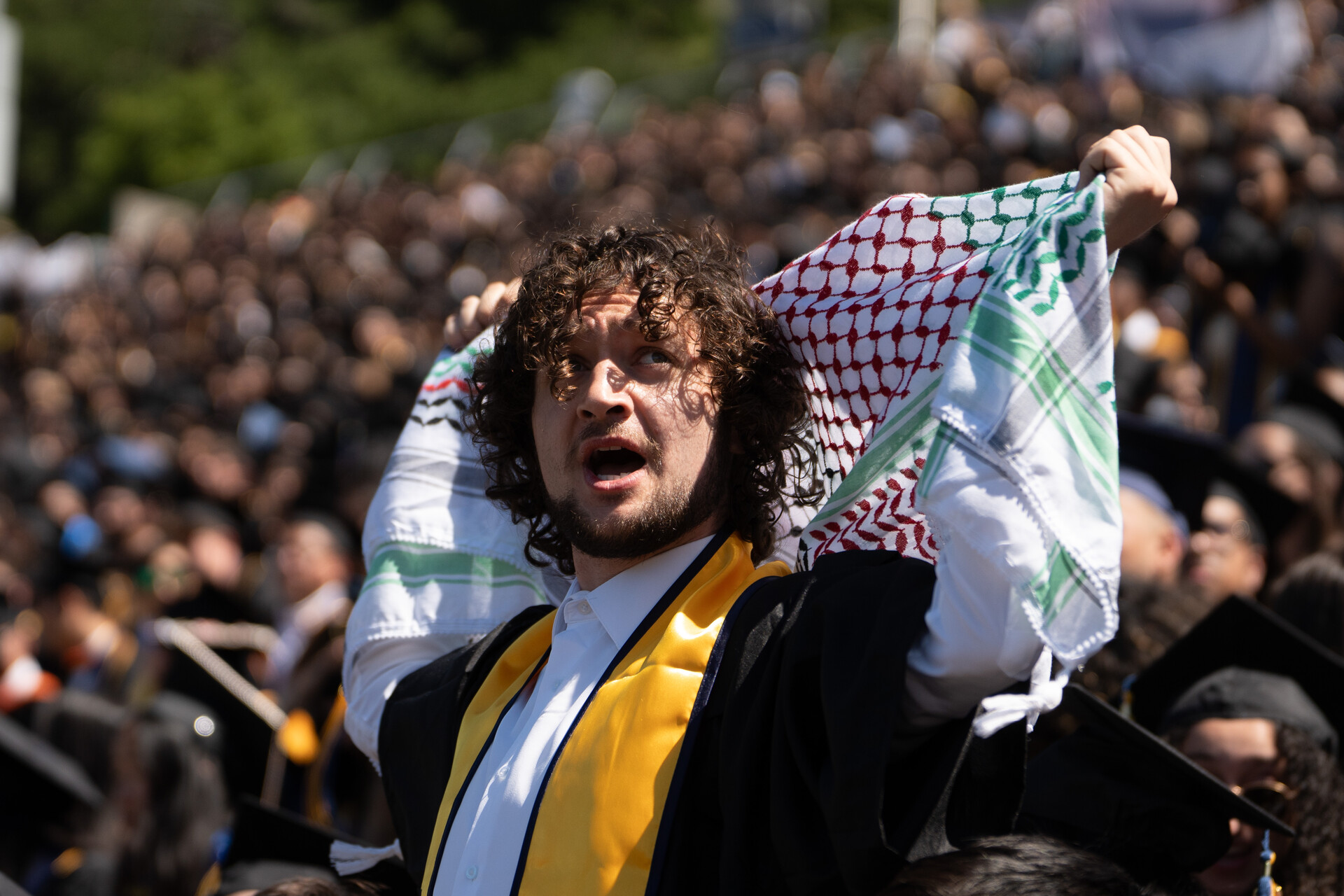A young man wearing a graduation gown holds a keffiyeh over his head with a crowd of other students in academic regalia behind him.
