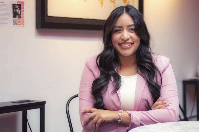 A Latina woman wearing a pink blazer seated with her arms crossed.