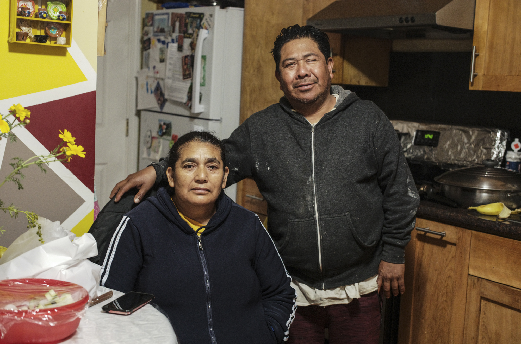A middle-aged Latinx couple, a woman seated and a man standing with his right arm around her as they both look at the camera in their home kitchen with a refrigerator behind them.